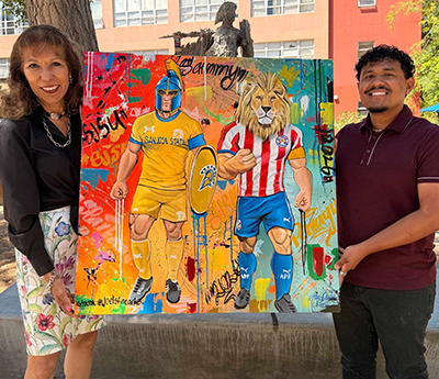 JSU President Cynthia Teniente-Matson and Joel Mendoza hold his piece depicting SJSU’s Spartan mascot, Sammy, and Team Paraguay’s lion mascot, Mbarte, in honor of the 2026 FIFA World Cup. 