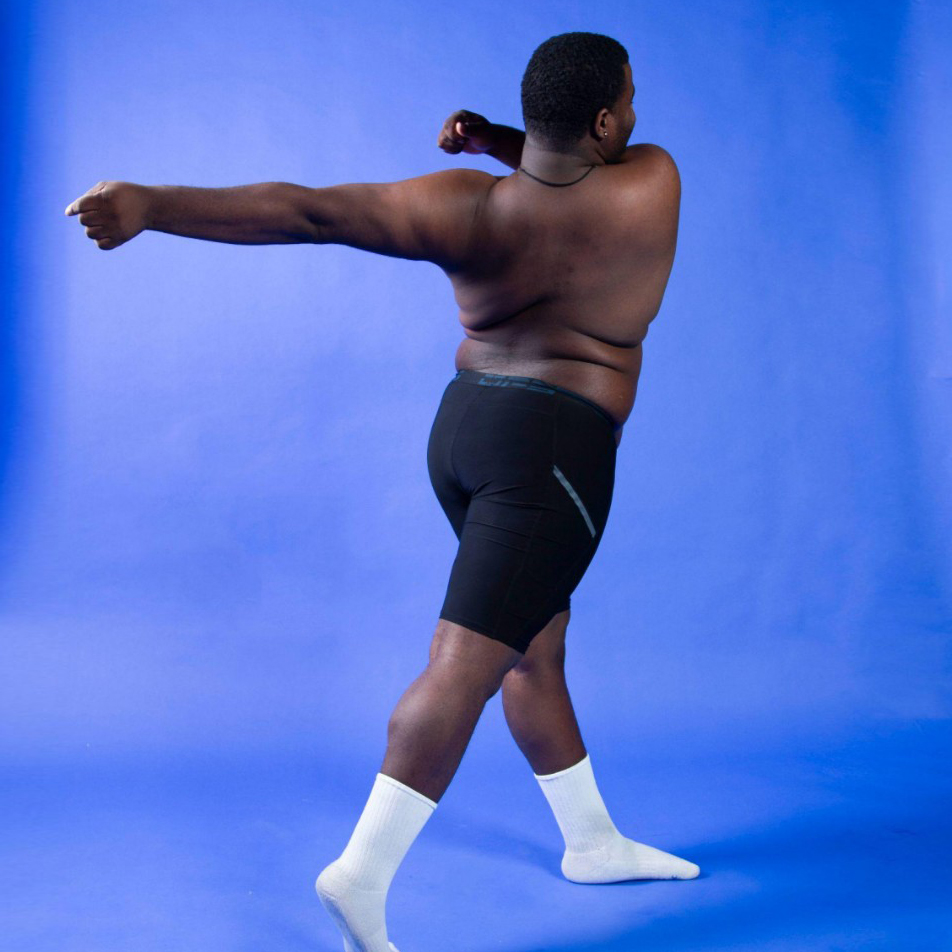 A person photographed from behind against a bright blue studio backdrop, standing in a dynamic pose with one arm extended to the side and the other bent near the shoulder. They are wearing black shorts and white socks, with their back turned slightly toward the camera.