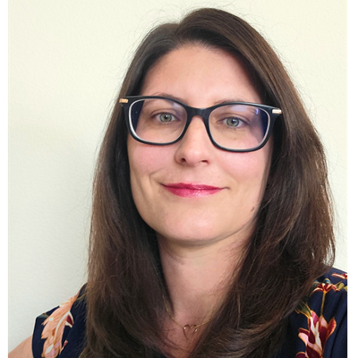 Self-portrait of Dominique Teaford, a pale-skinned woman with brunette hair, wearing a black and white blazer. In the background is an outdoor space with trees.