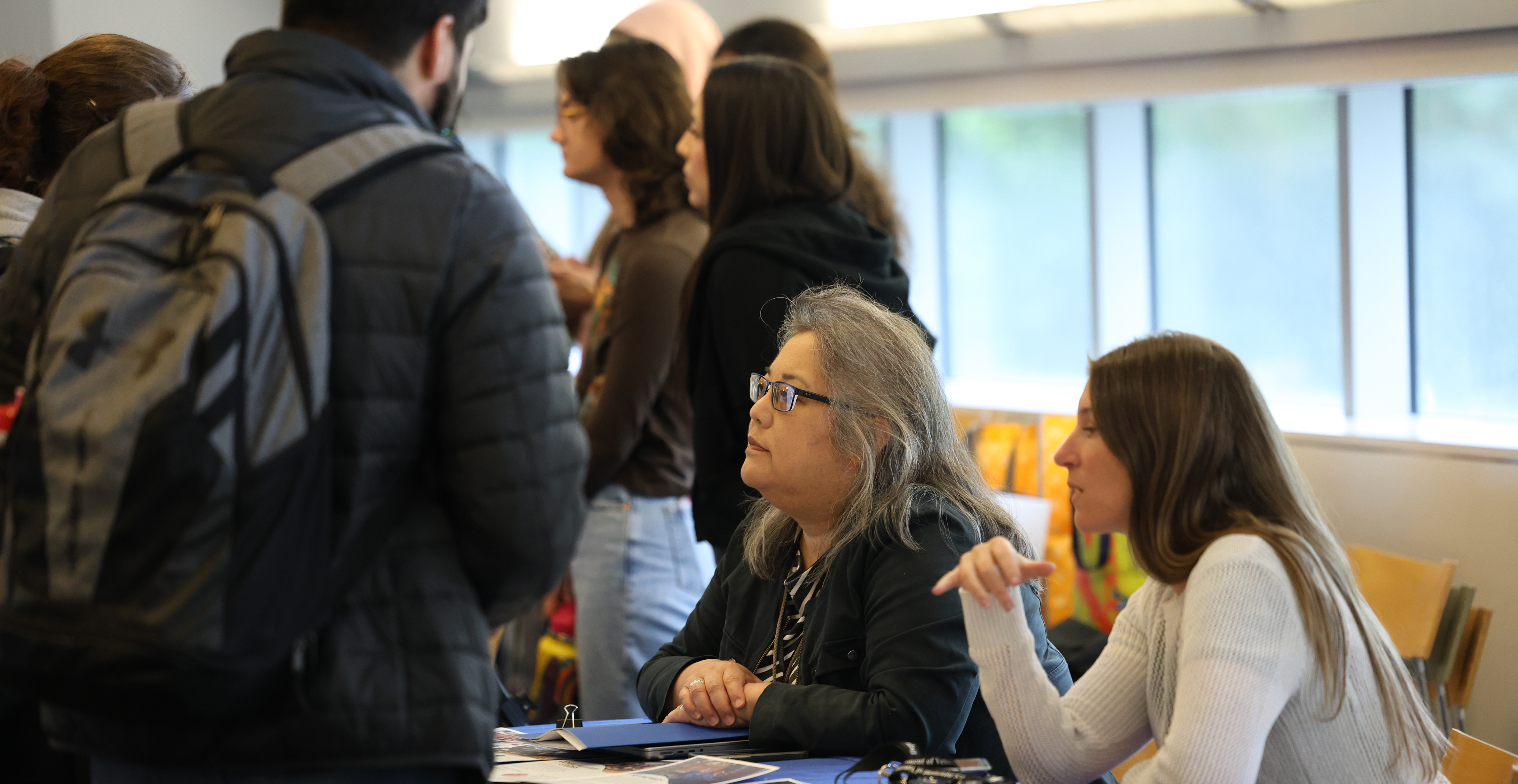 A professor and staff member or student assistant sit at table full of materials, talking to students. More tablers and students are in back.